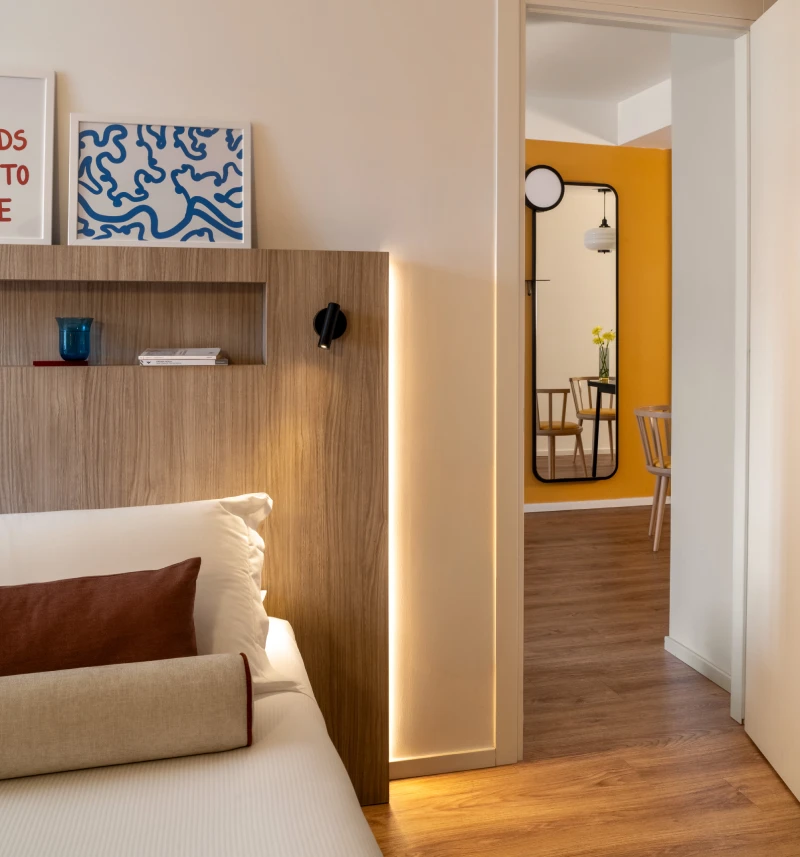 Room detail with bed, wood headboard, warm lighting and access to a yellow living area - Aparthotel Colombo, BZAR hotels in Rome